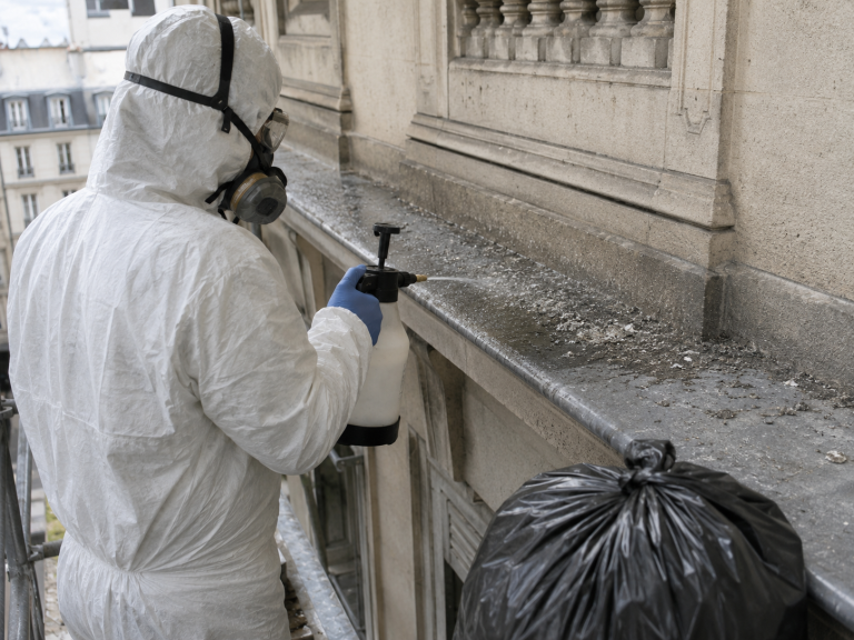 Opérateur en combinaison et masque respiratoire humidifiant une corniche souillée au pulvérisateur